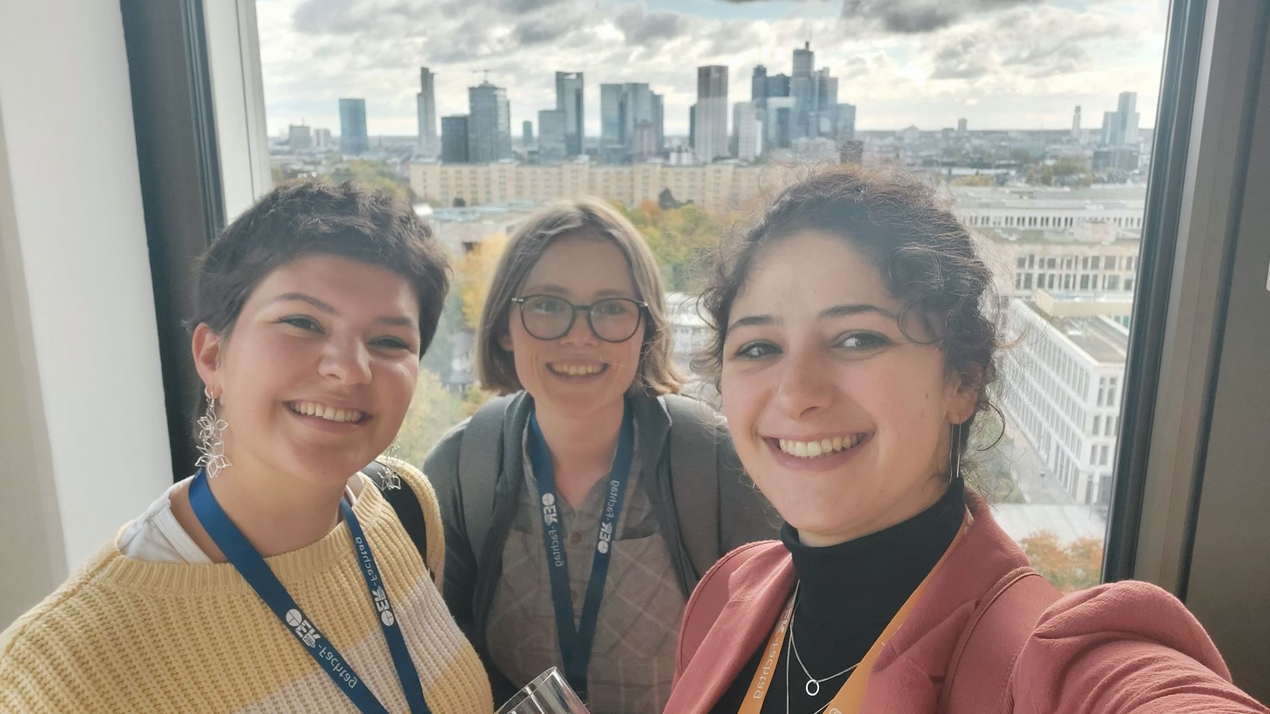 PrimOER vernetzt sich mit den Projekten KlimaOER und digillcom. Auf dem Bild sind 3 Vertreterinnnen der jeweiligen Projekte zu sehen, die gemeinsam in die Kamera lächeln. Im Hintergrund ist die Skyline von Frankfurt zu sehen.